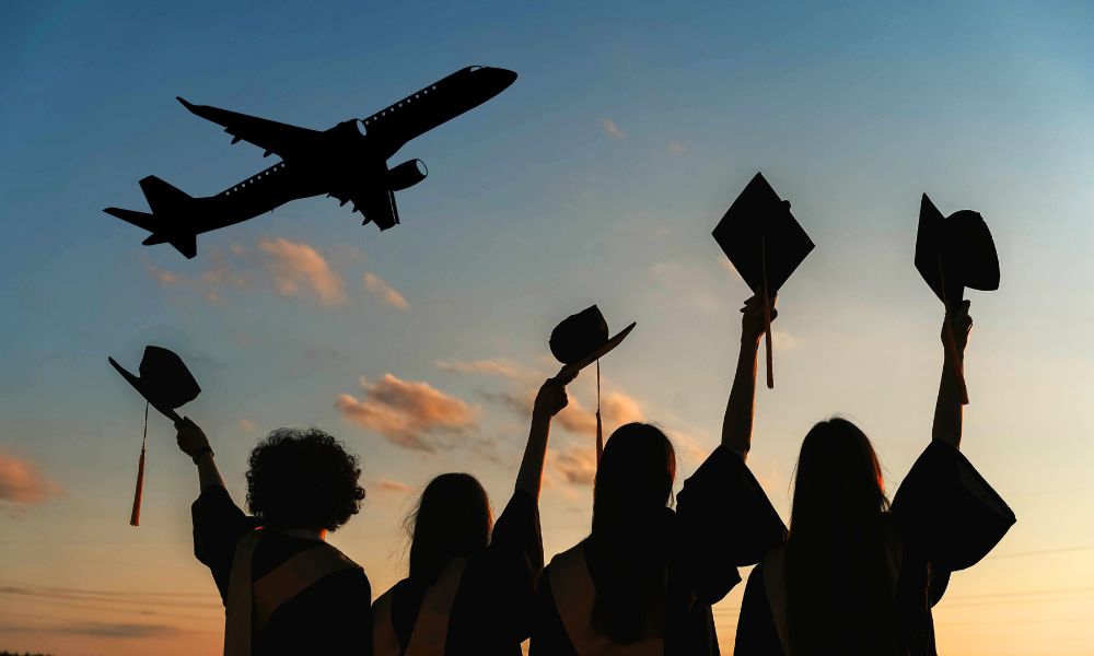 Silhouette of students waving their graduation caps to an overhead airplane at sunset